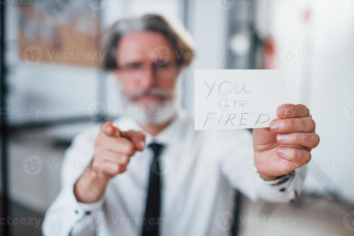 Holding paper with text you are fired. Mature businessman with grey hair and beard in formal clothes is in the office photo