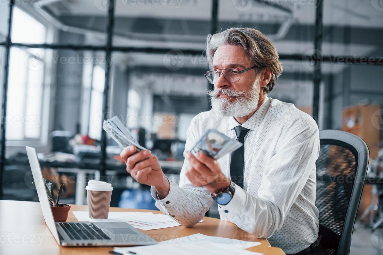 Happy mature businessman with grey hair and beard in formal clothes is in the office holding money photo