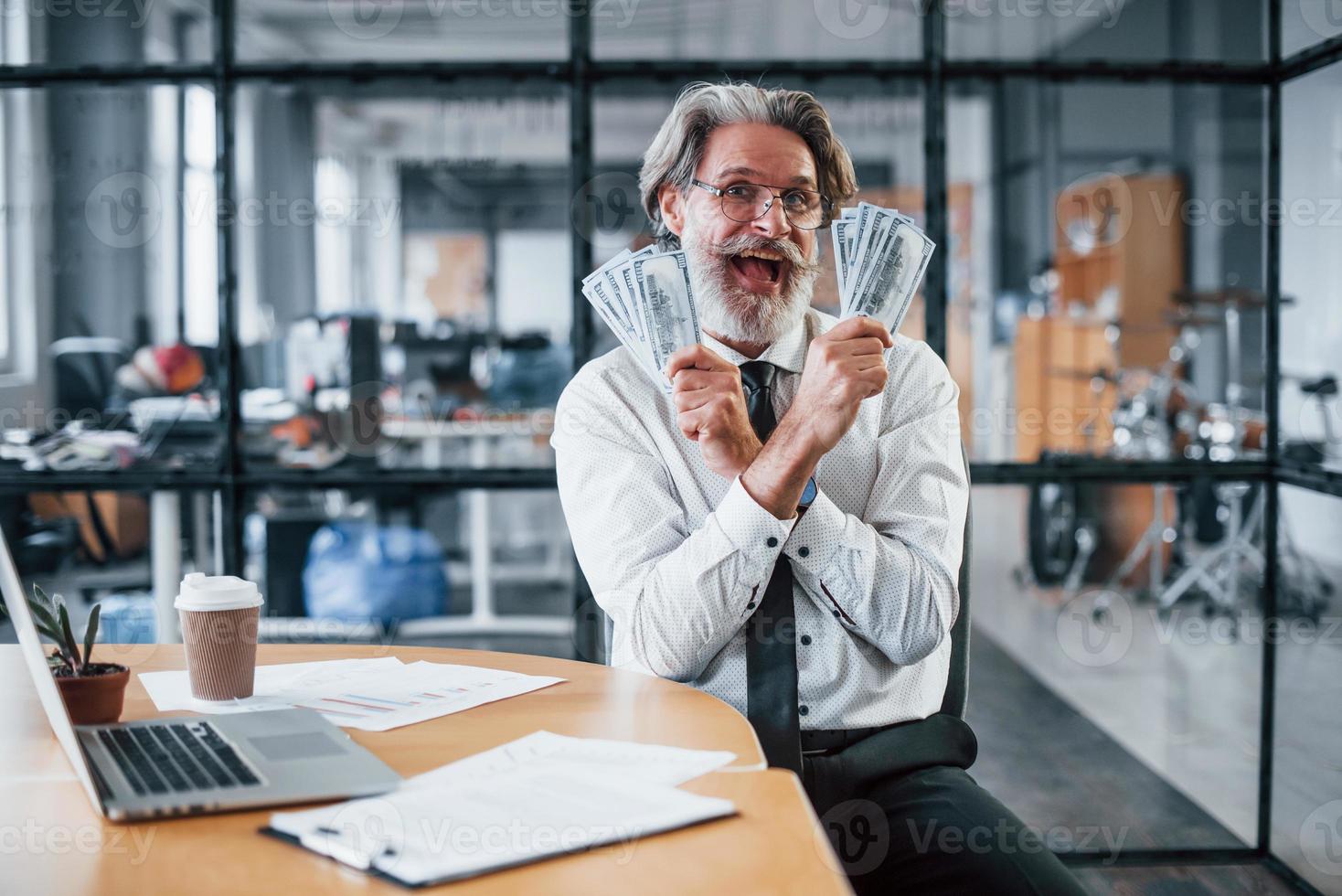 Happy mature businessman with grey hair and beard in formal clothes is in the office holding money photo