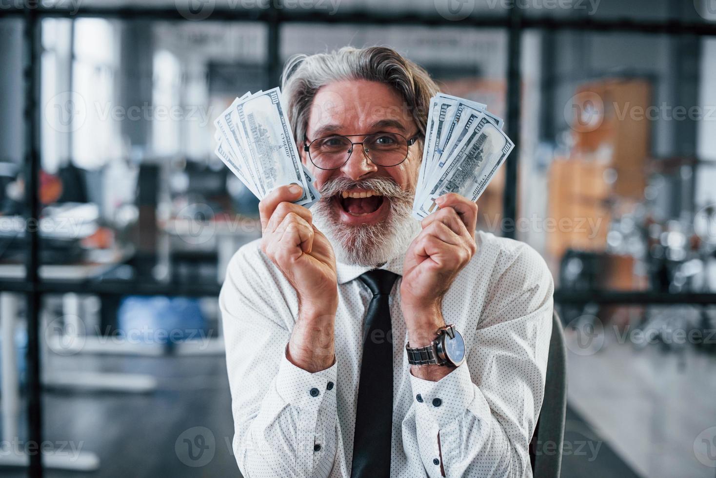 Happy mature businessman with grey hair and beard in formal clothes is in the office holding money photo