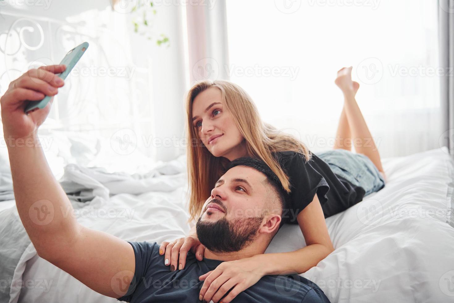 joven pareja casada haciendo un selfie usando el teléfono en el dormitorio durante el día foto