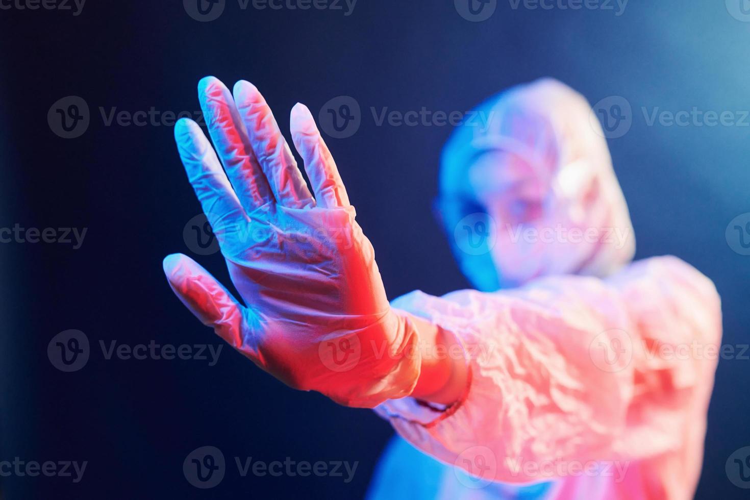 enfermera con máscara y uniforme blanco parada en una habitación iluminada con neón y mostrando la señal de alto foto