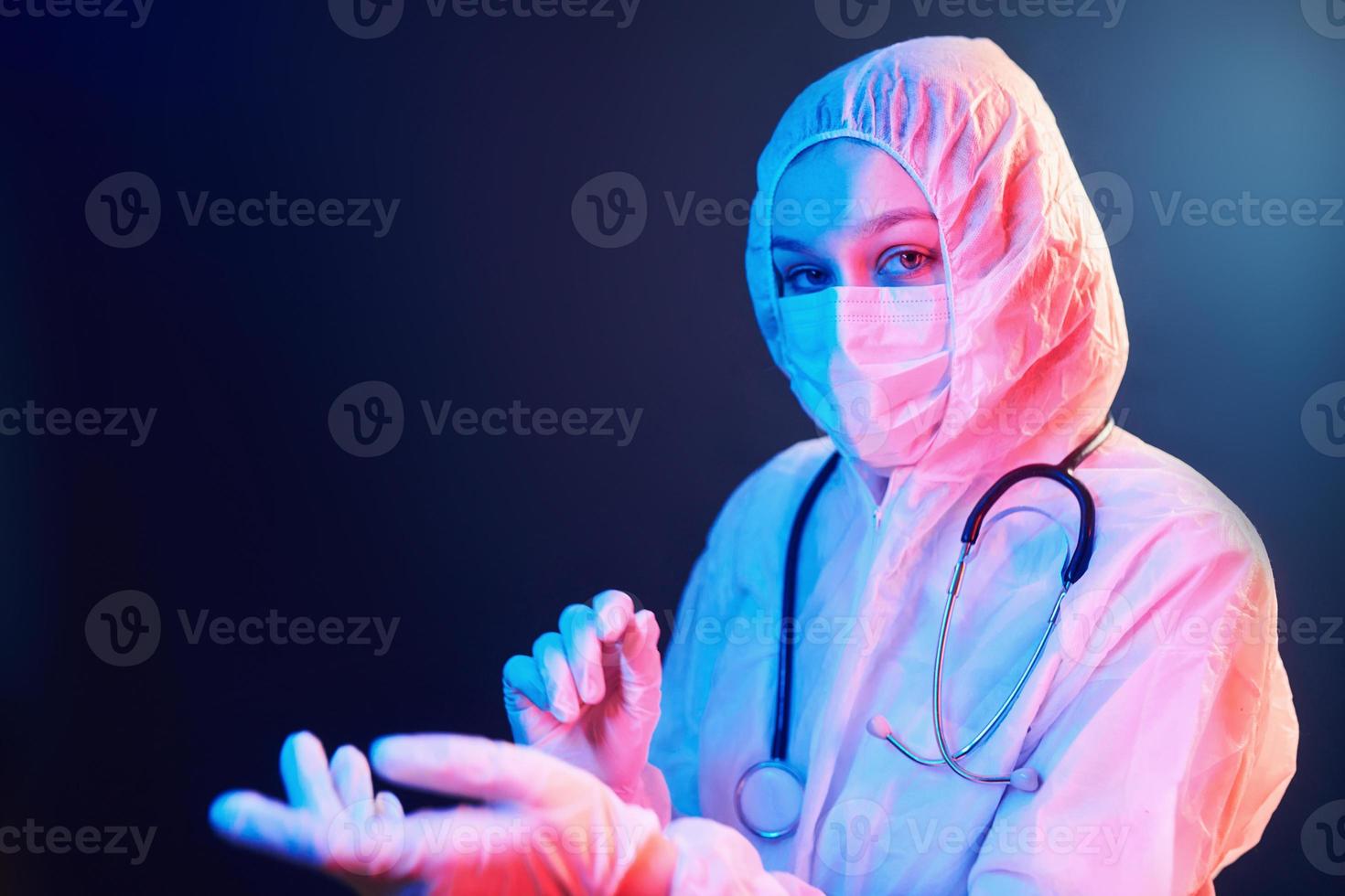 Nurse in mask and white uniform and with stethoscope standing in neon lighted room photo