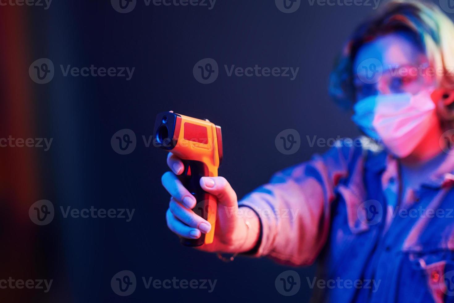 Girl in protective glasses and mask holding infrared thermometer. Neon lights photo