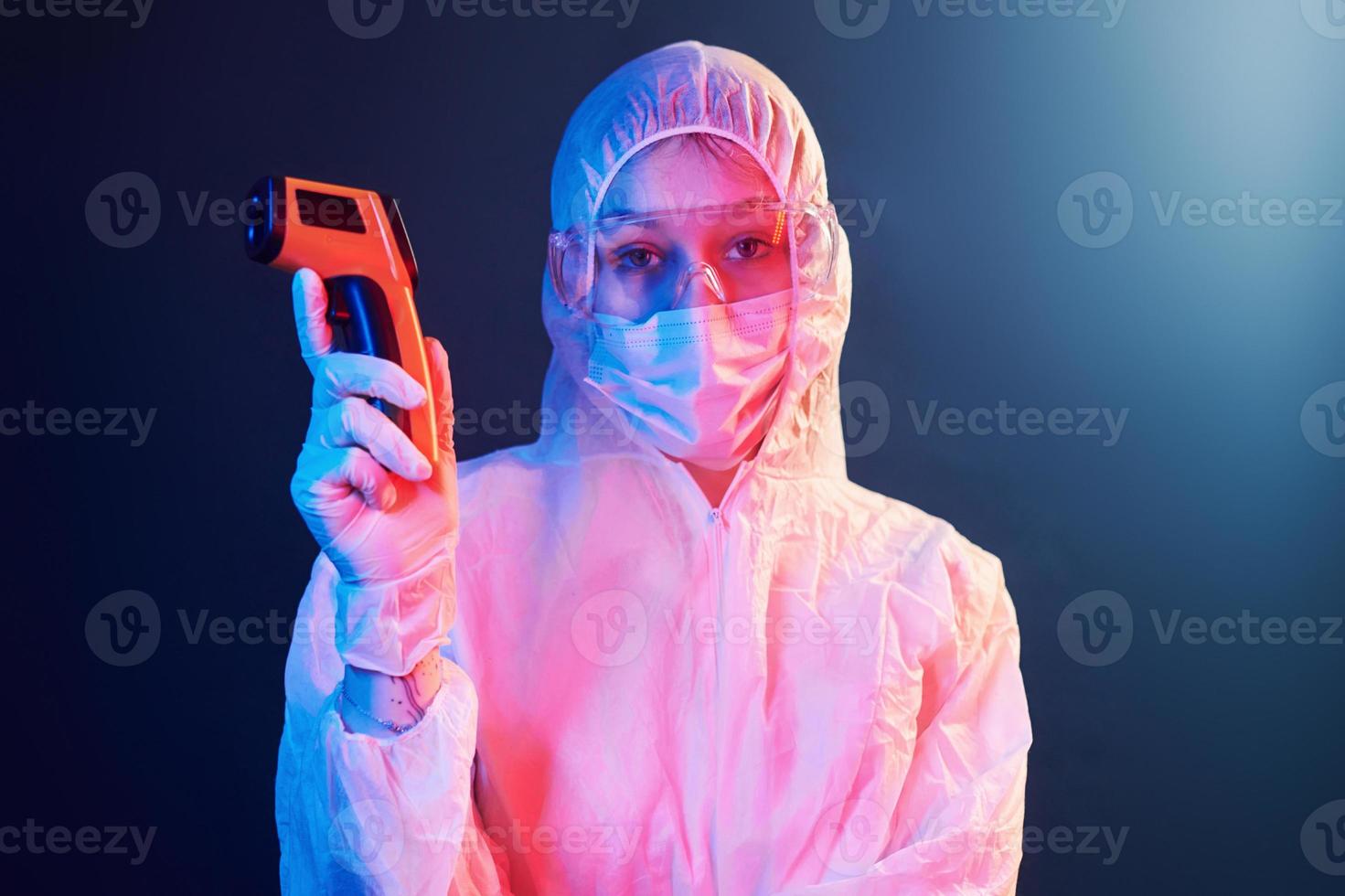 Nurse in mask and white uniform standing in neon lighted room and holding infrared thermometer. Stop coronavirus spreading photo