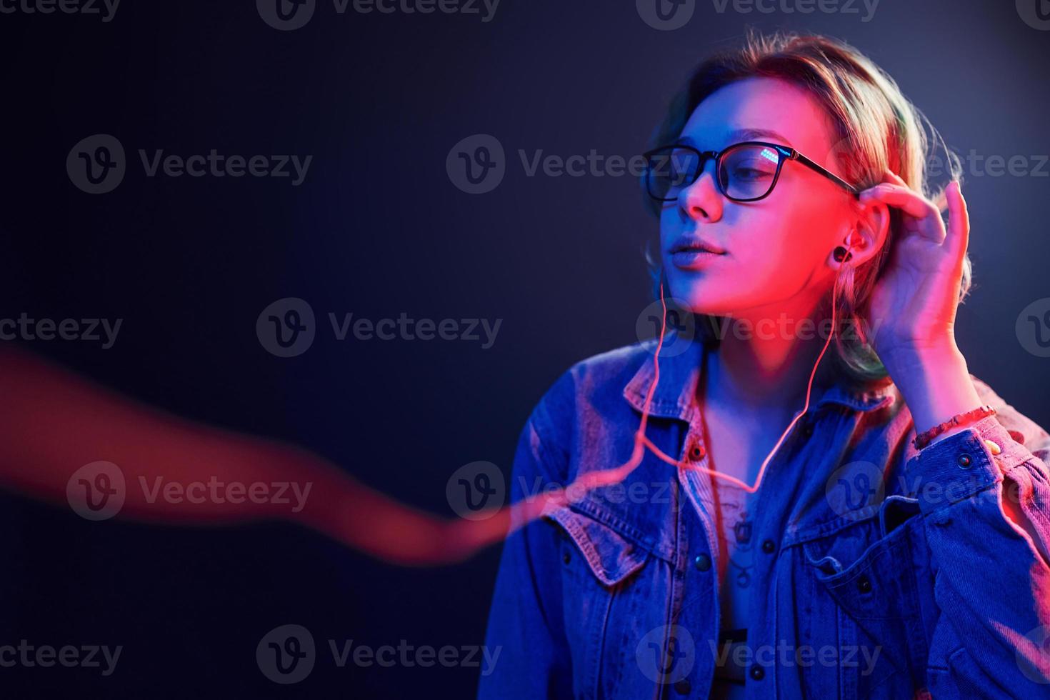 Portrait of young alternative girl in glasses with green hair in red and blue neon light in studio that listening to music in headphones photo
