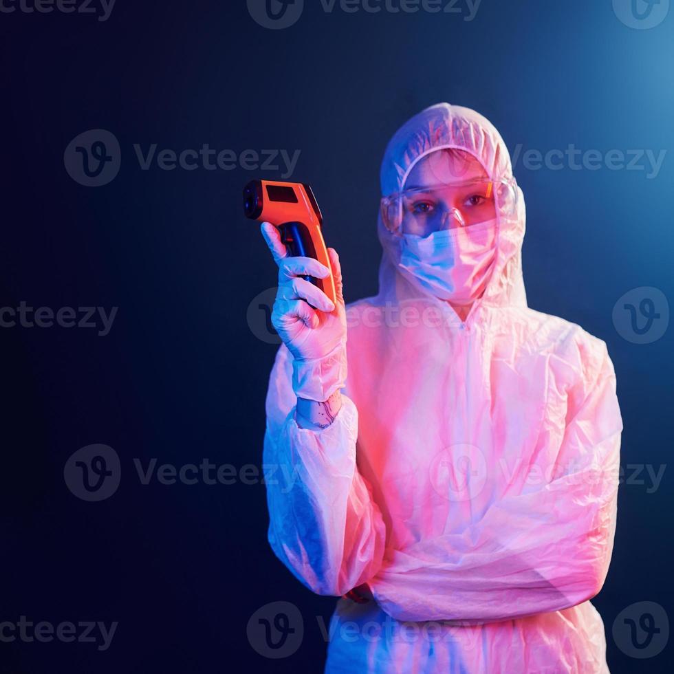 enfermera con máscara y uniforme blanco parada en una habitación iluminada con neón y sosteniendo un termómetro infrarrojo. detener la propagación del coronavirus foto