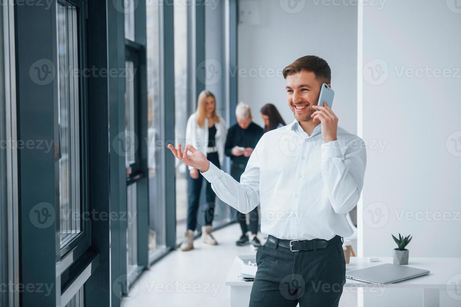 Man in formal clothes talking by phone and standing in front of young successful team that working and communicating together indoors in office photo