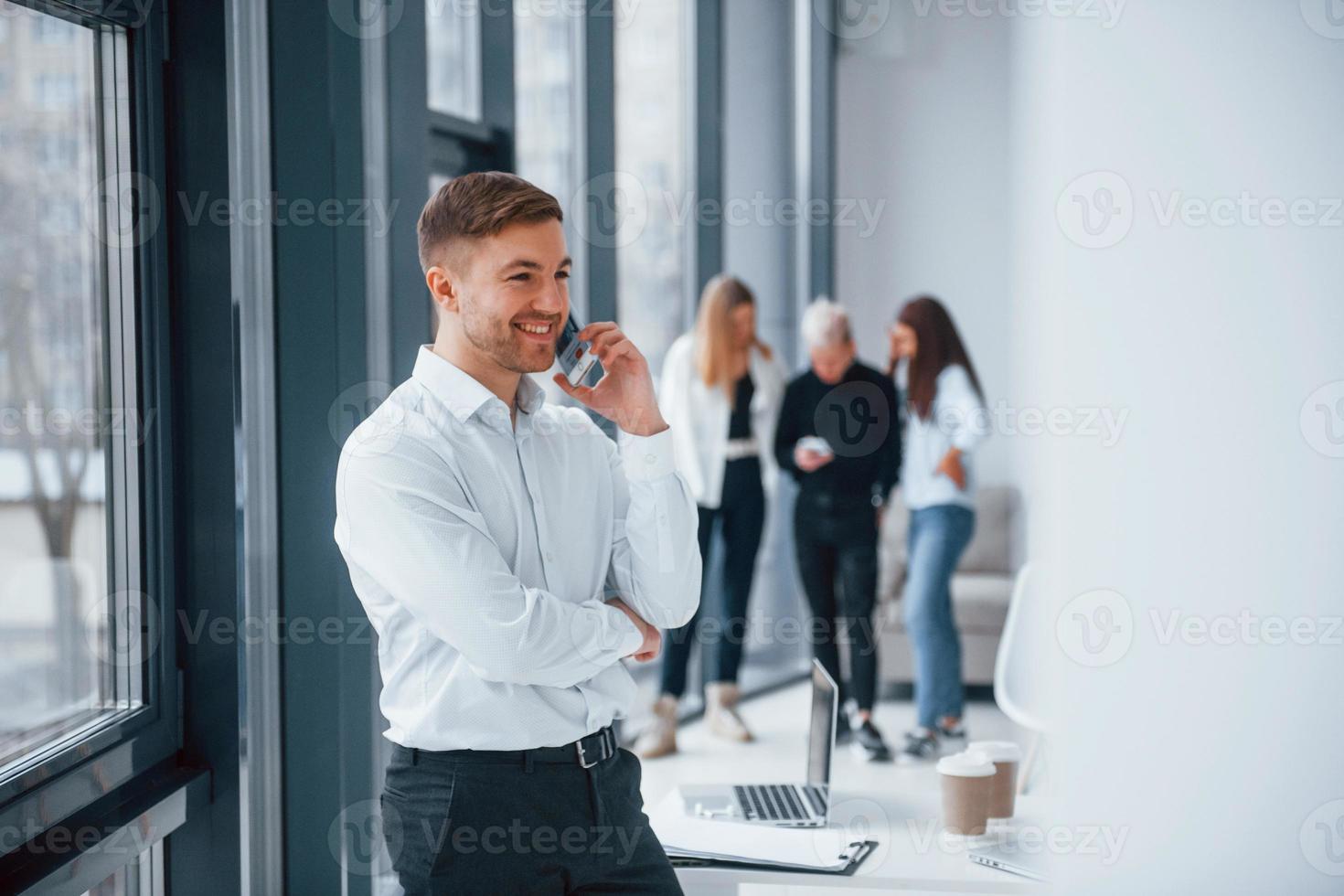 Man in formal clothes talking by phone and standing in front of young successful team that working and communicating together indoors in office photo