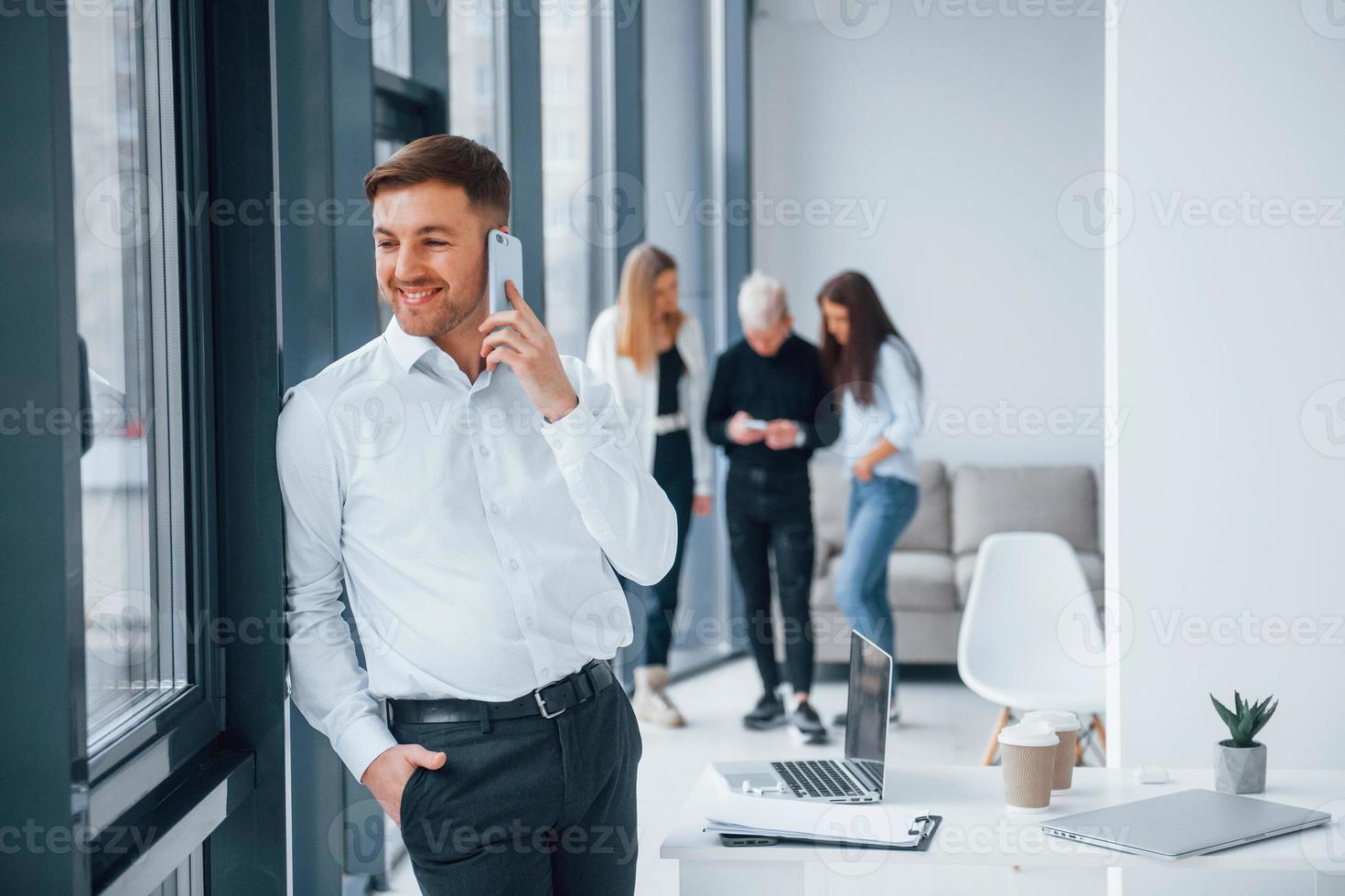 Man in formal clothes talking by phone and standing in front of young successful team that working and communicating together indoors in office photo
