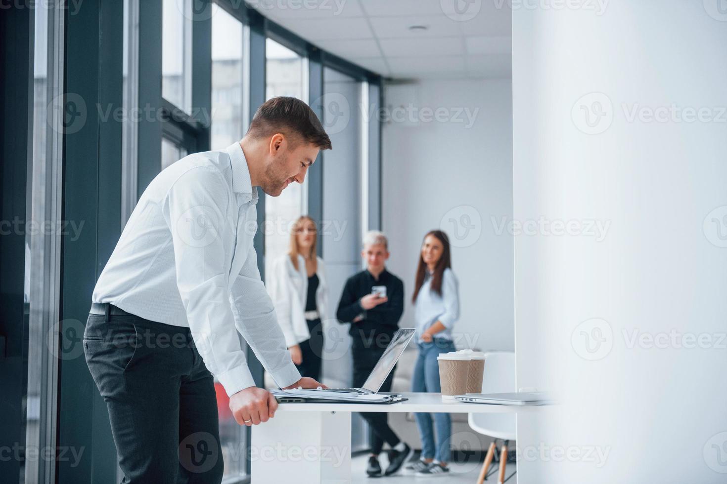 Man in formal clothes standing and reading documents in front of young successful team that working and communicating together indoors in office photo