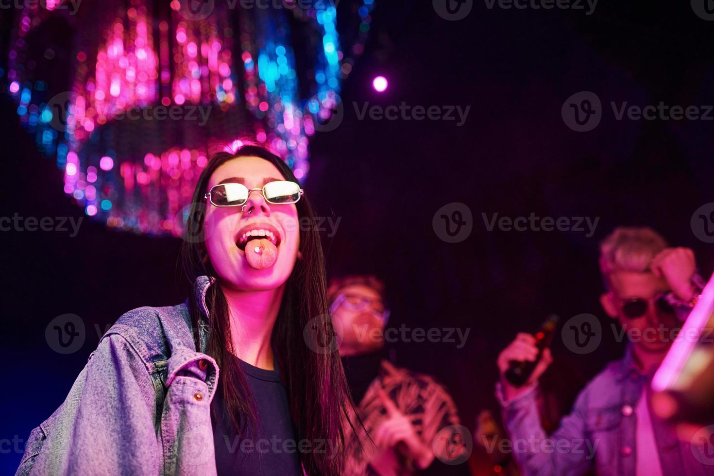 niña sosteniendo pastillas de drogas en togue frente a los jóvenes que se divierten en el club nocturno con luces láser de colores foto