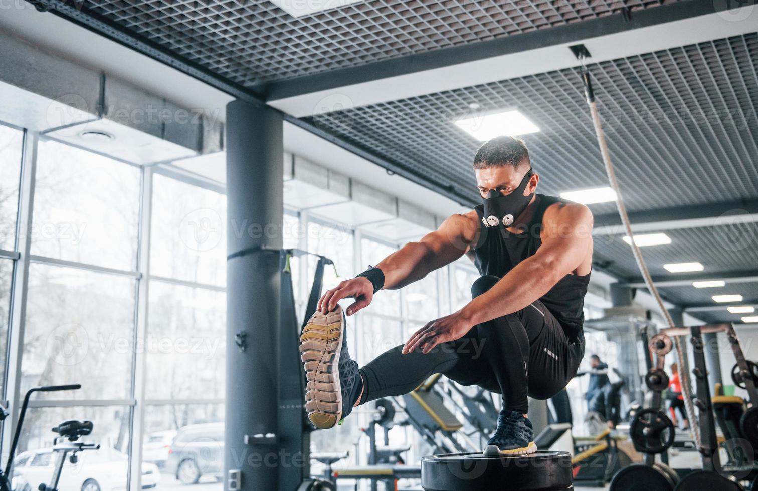 Strong young man in sportive clothes and scary mask doing push ups on one leg in gym photo