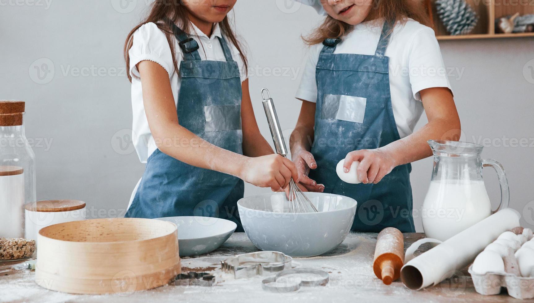 dos niñas pequeñas con uniforme de chef azul mezclando harina con huevos en un plato en la cocina foto