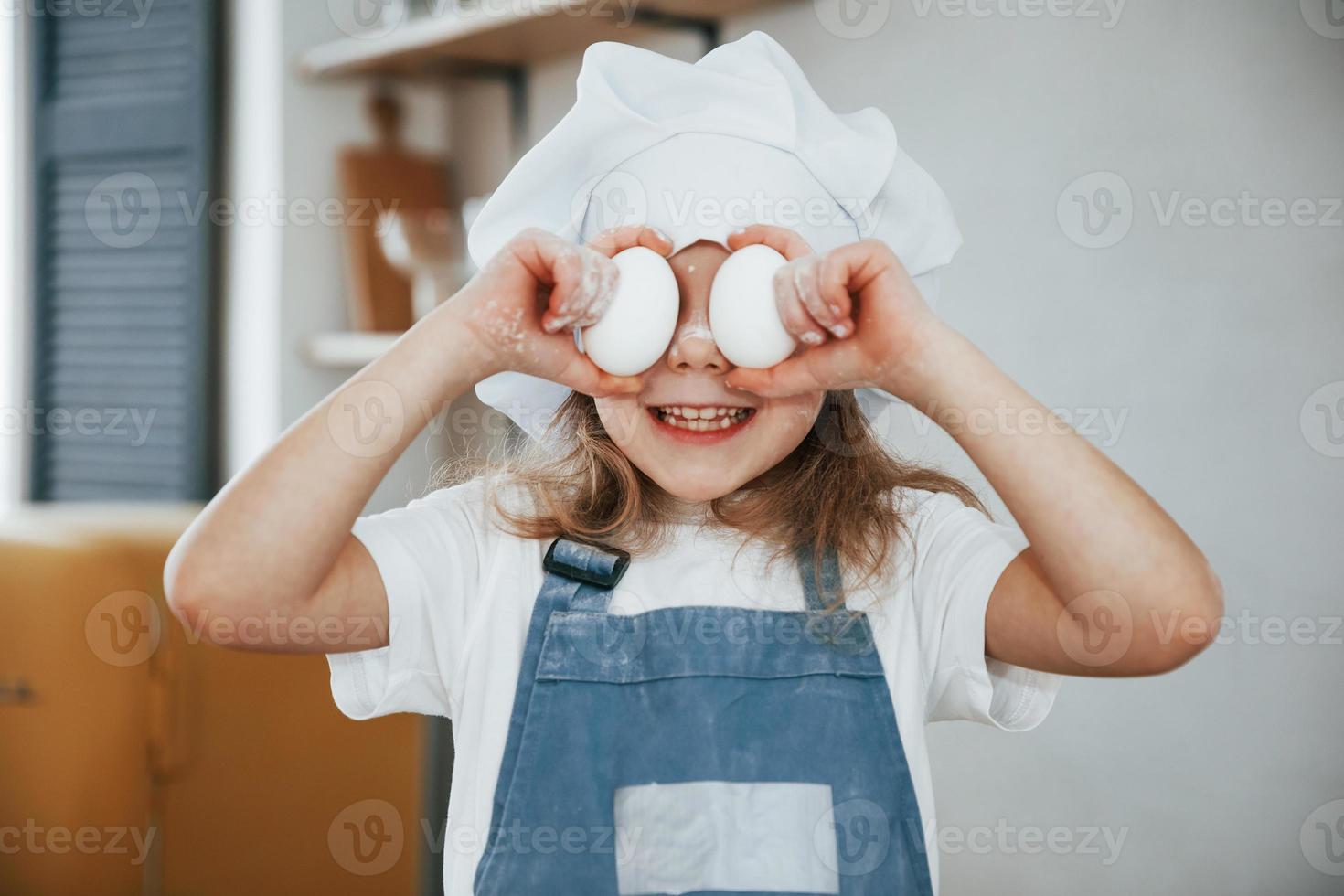 niñita alegre con sombrero blanco y uniforme de chef azul sonriendo y cubriendo sus ojos con huevos foto