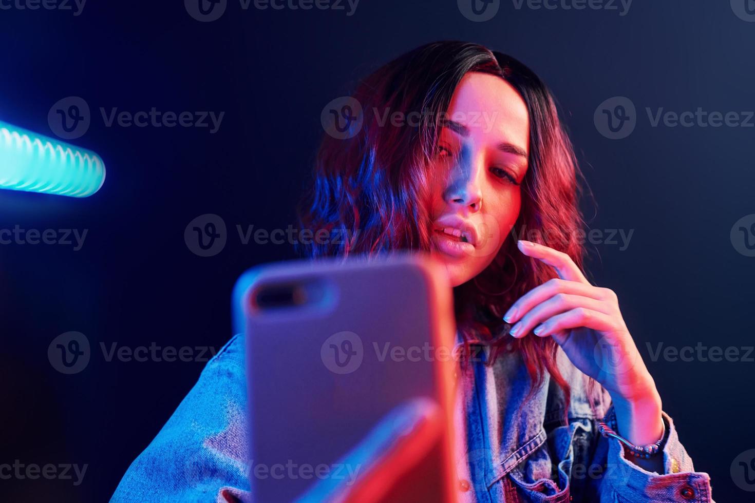Portrait of young girl with curly hair that makes selfie by using phone in red and blue neon in studio photo