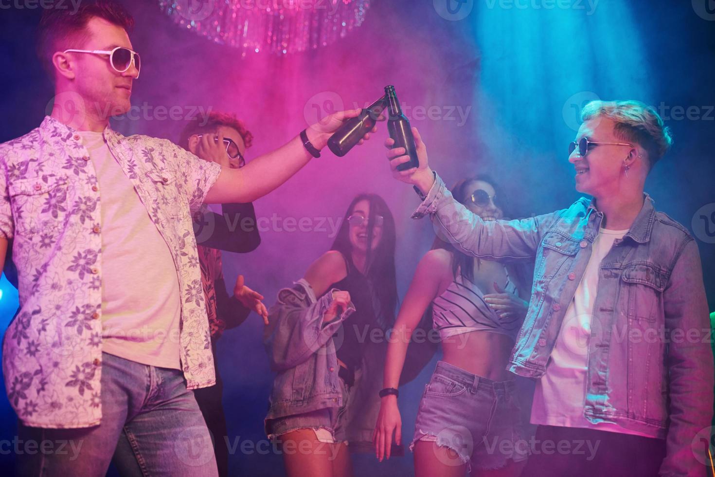 Celebrating and knocking bottles with alcohol. Young people is having fun in night club with colorful laser lights photo