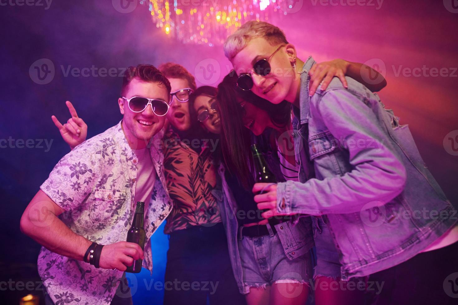 With bottles of beer in hands. Young people is having fun in night club with colorful laser lights photo