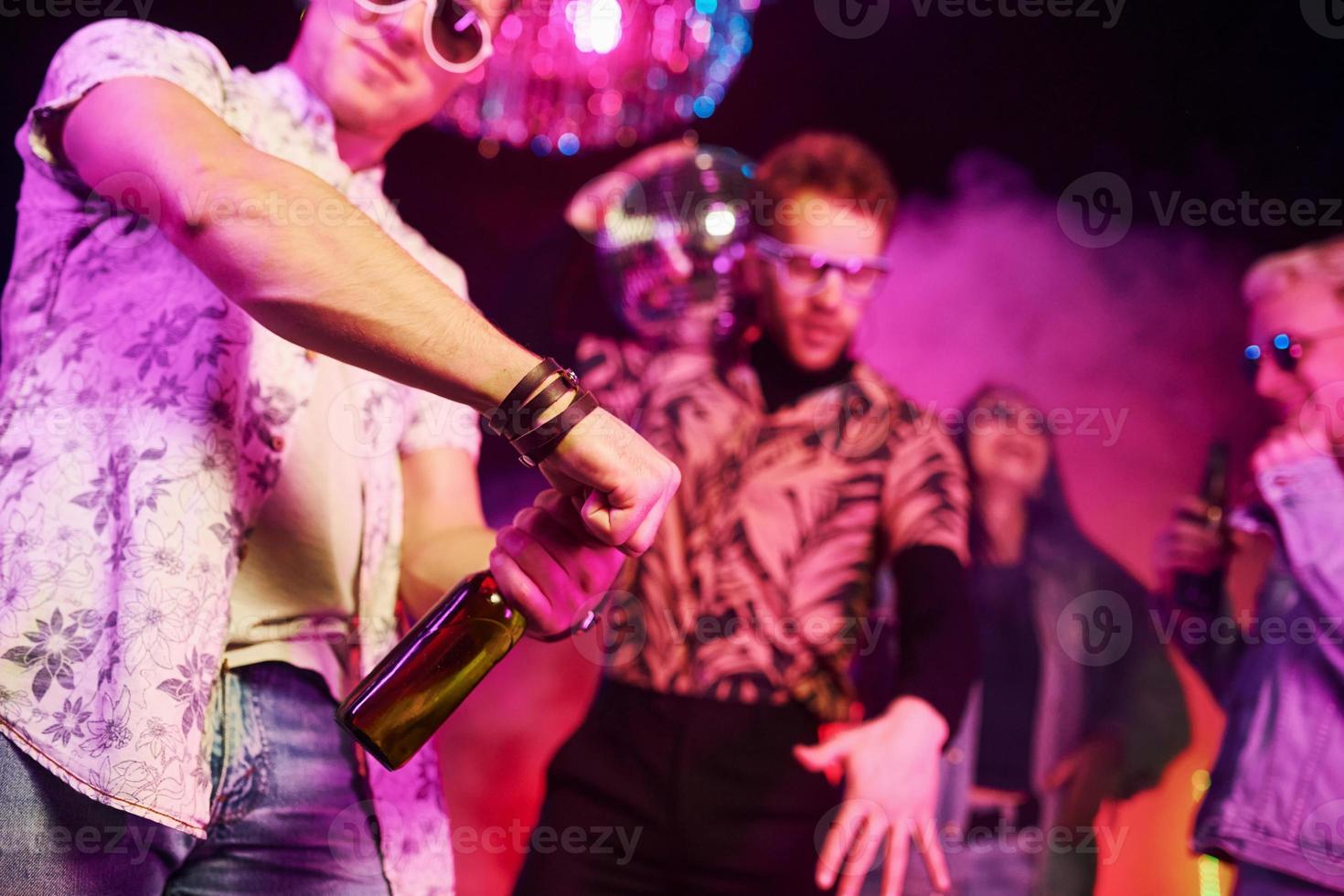 Celebrating and knocking bottles with alcohol. Young people is having fun in night club with colorful laser lights photo