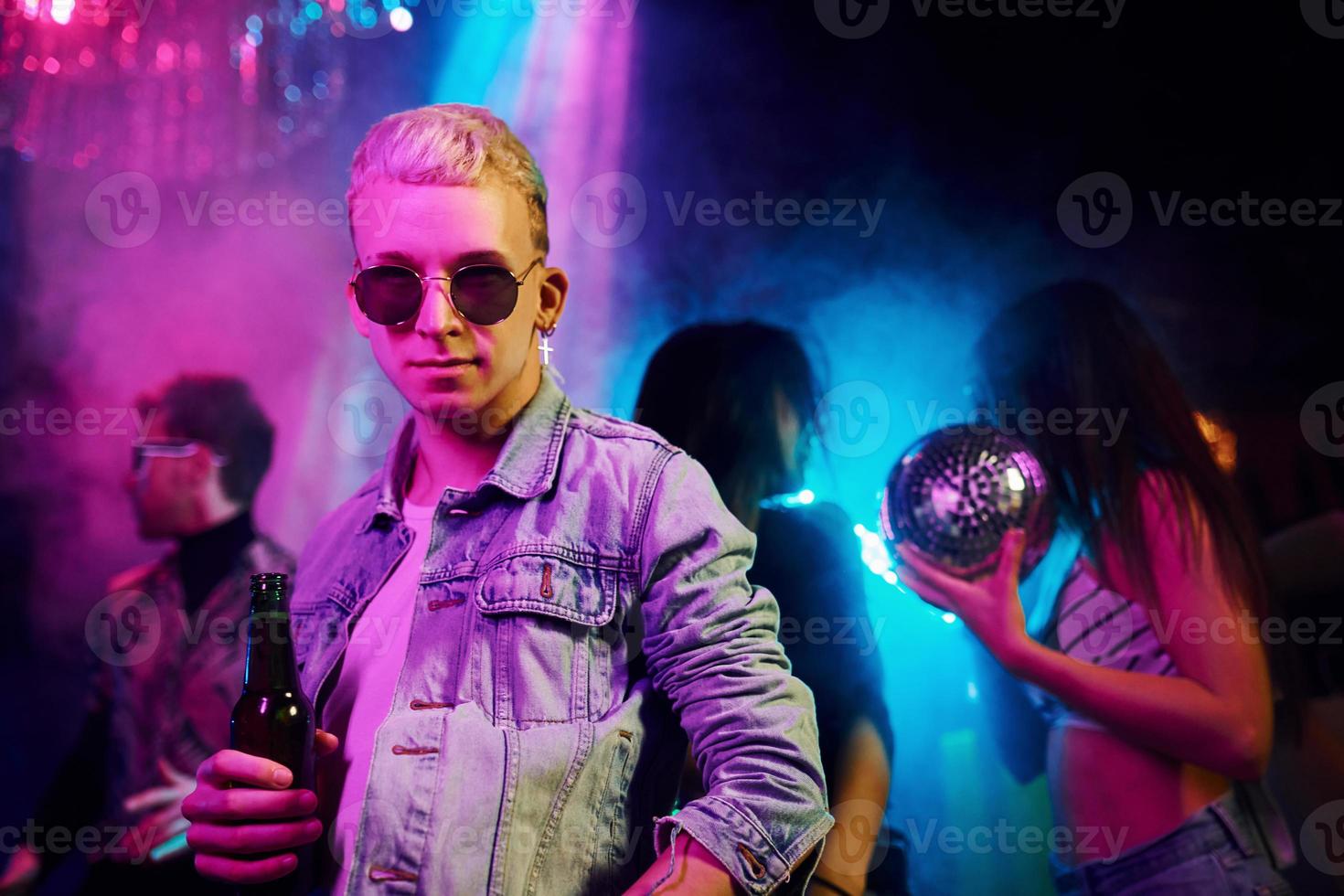 Hipster guy in sunglasses and with bottle of alcohol posing for camera in front of young people that having fun in night club with colorful laser lights photo