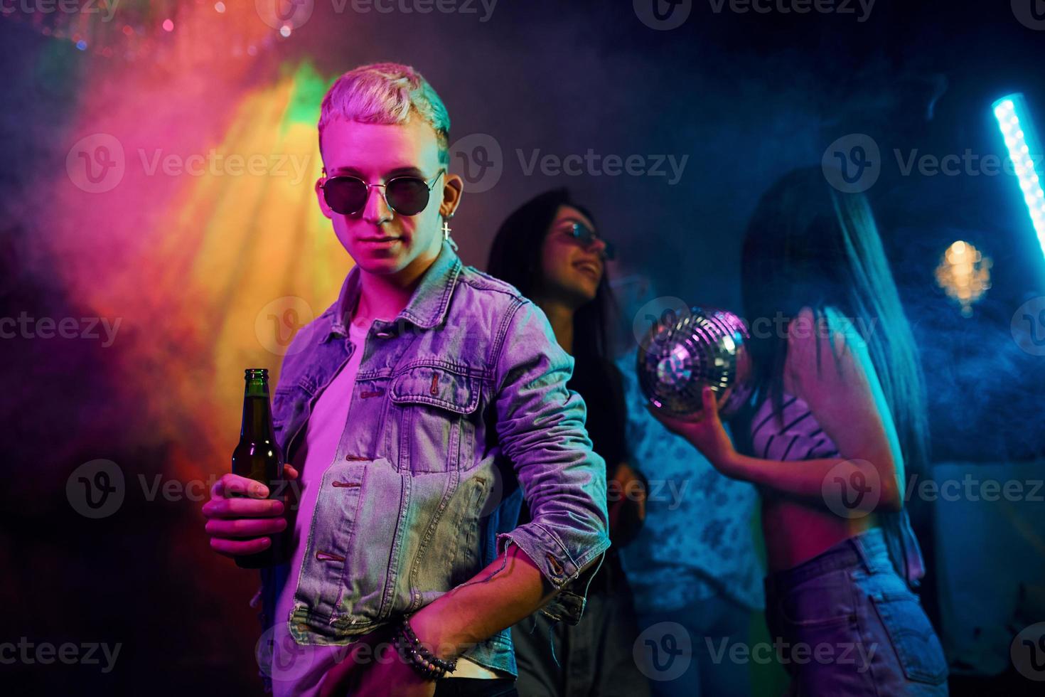 Hipster guy in sunglasses and with bottle of alcohol posing for camera in front of young people that having fun in night club with colorful laser lights photo