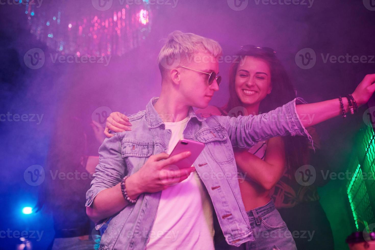 chico parado con un teléfono inteligente frente a jóvenes que se divierten en un club nocturno con luces láser de colores foto