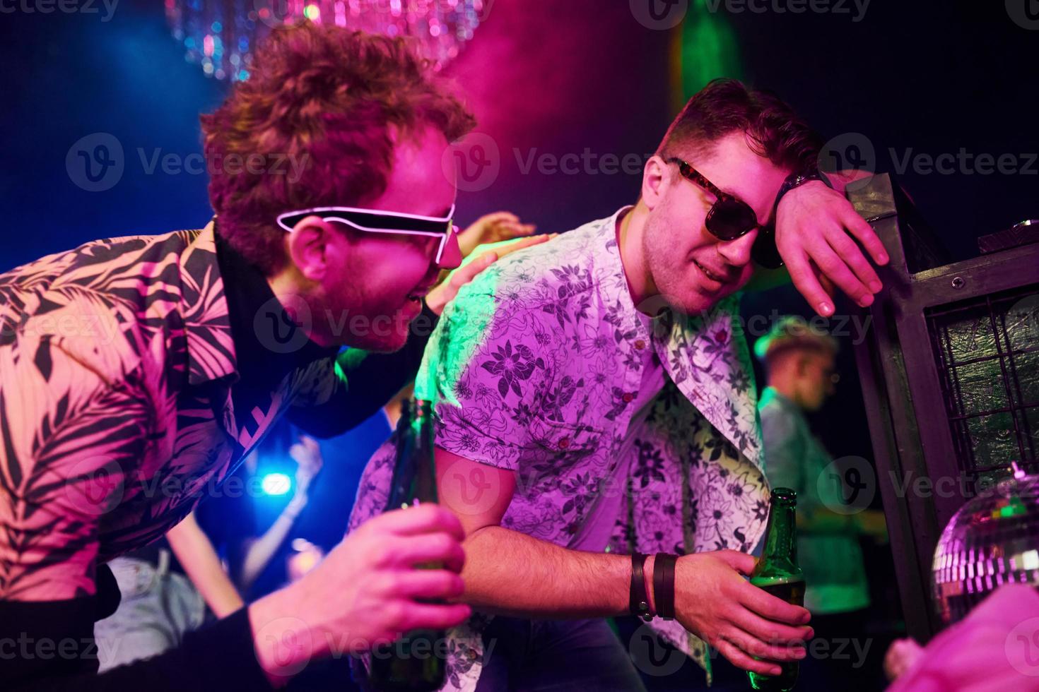 borracho apoyado en la cerca frente a los jóvenes que se divierten en el club nocturno con luces láser de colores foto