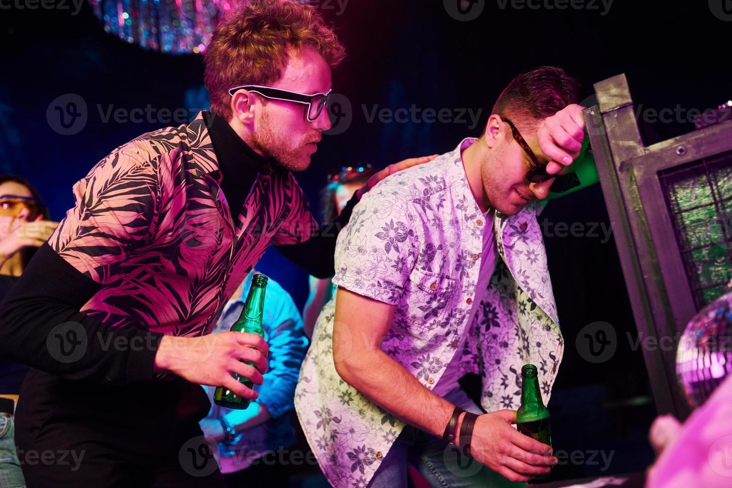 Drunk guy leaning in fence in front of young people that having fun in night club with colorful laser lights photo