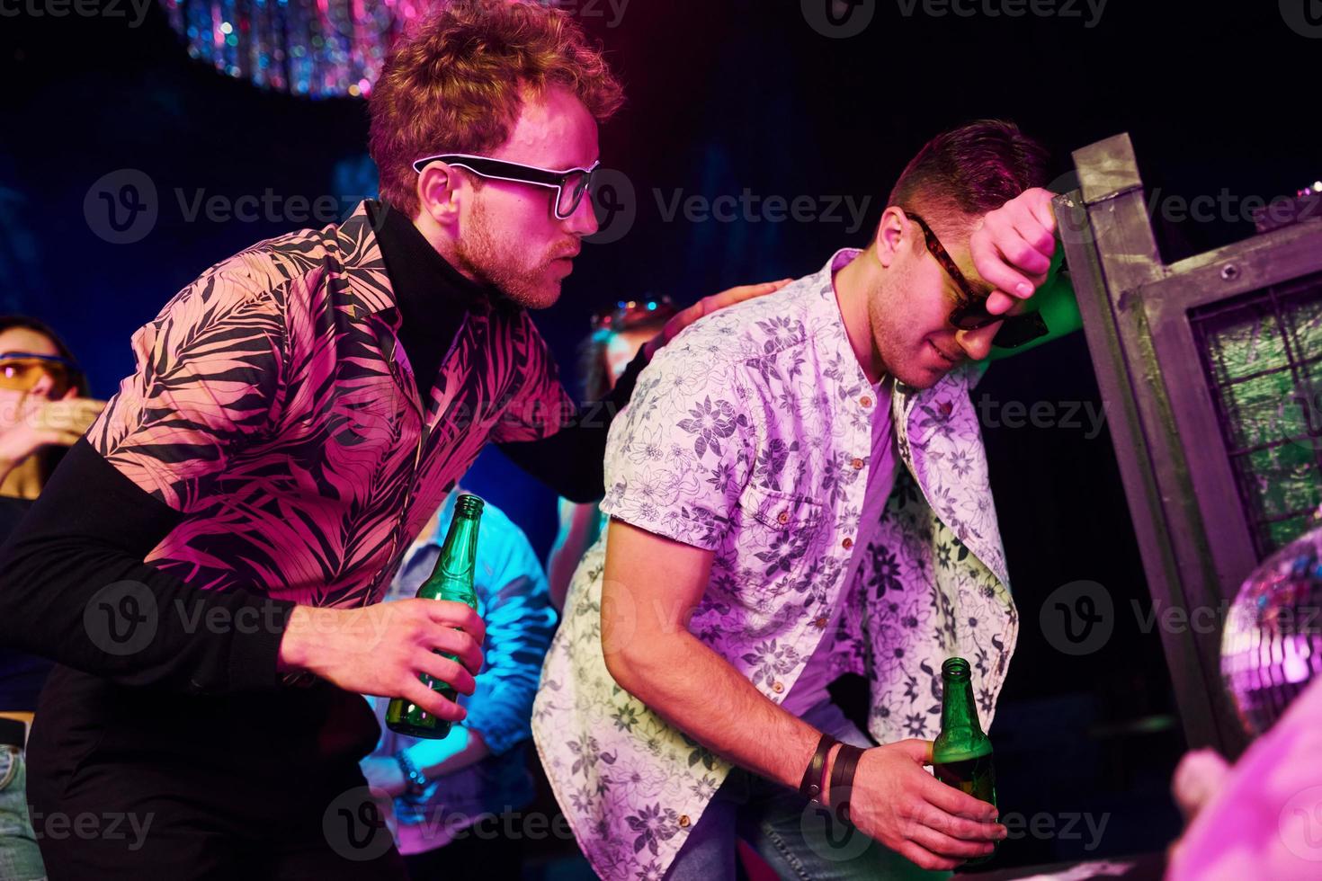 borracho apoyado en la cerca frente a los jóvenes que se divierten en el club nocturno con luces láser de colores foto