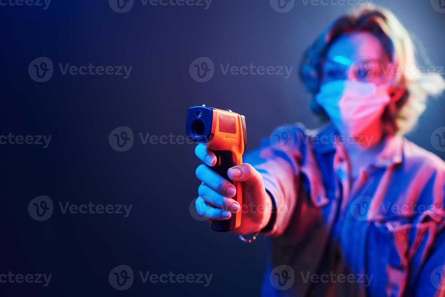 Girl in protective glasses and mask holding infrared thermometer. Neon lights photo
