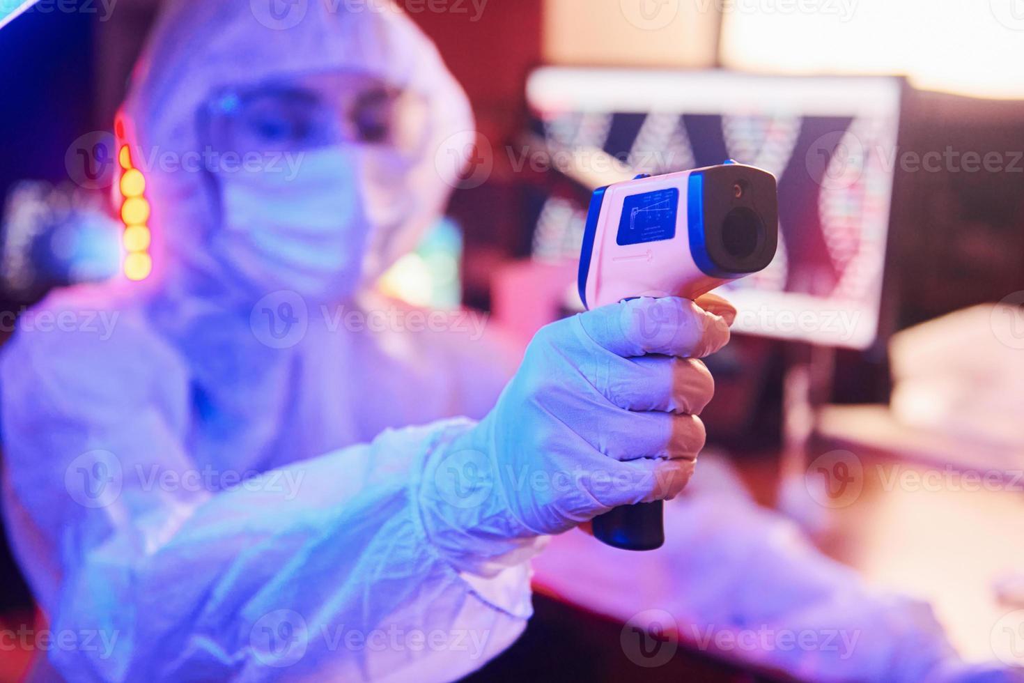 Nurse in mask and white uniform, holding infrared thermometer and sitting in neon lighted laboratory with computer and medical equipment searching for Coronavirus vaccine photo