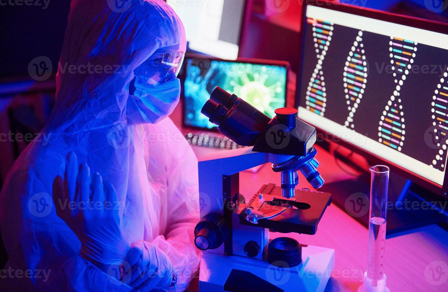 Nurse in mask and white uniform sitting in neon lighted laboratory with computer, microscope and medical equipment searching for Coronavirus vaccine photo