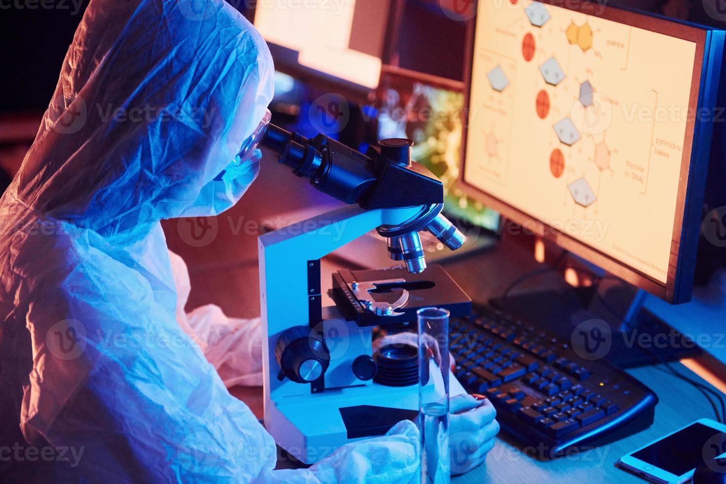 Nurse in mask and white uniform sitting in neon lighted laboratory with computer, microscope and medical equipment searching for Coronavirus vaccine photo
