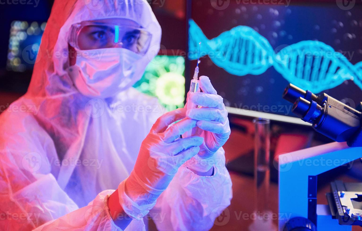 enfermera con máscara y uniforme blanco, sosteniendo una jeringa y sentada en un laboratorio iluminado con neón con computadora y equipo médico en busca de la vacuna contra el coronavirus foto