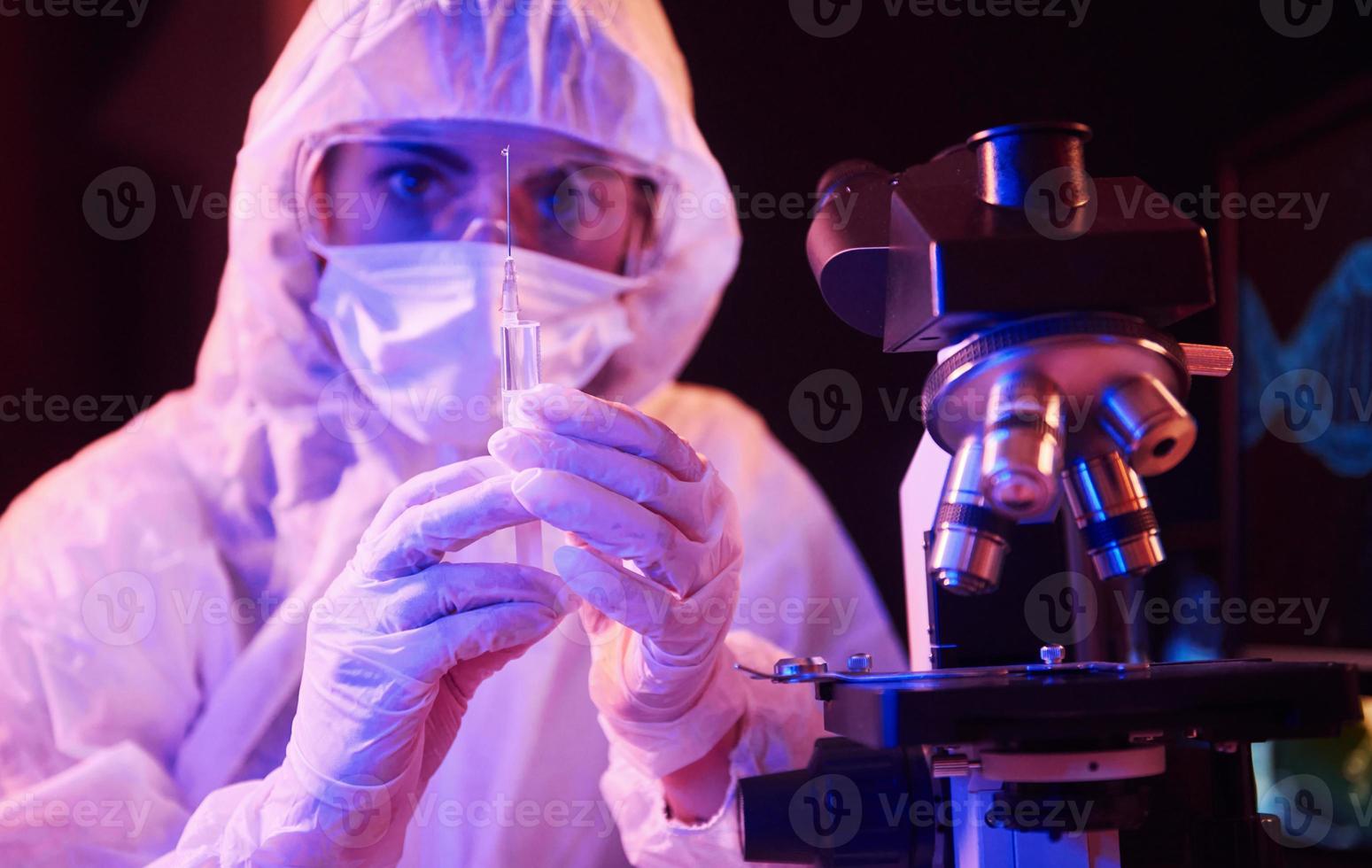 enfermera con máscara y uniforme blanco, sosteniendo una jeringa y sentada en un laboratorio iluminado con neón con computadora y equipo médico en busca de la vacuna contra el coronavirus foto