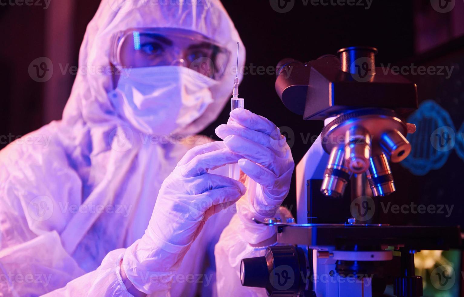 Nurse in mask and white uniform, holding syringe and sitting in neon lighted laboratory with computer and medical equipment searching for Coronavirus vaccine photo