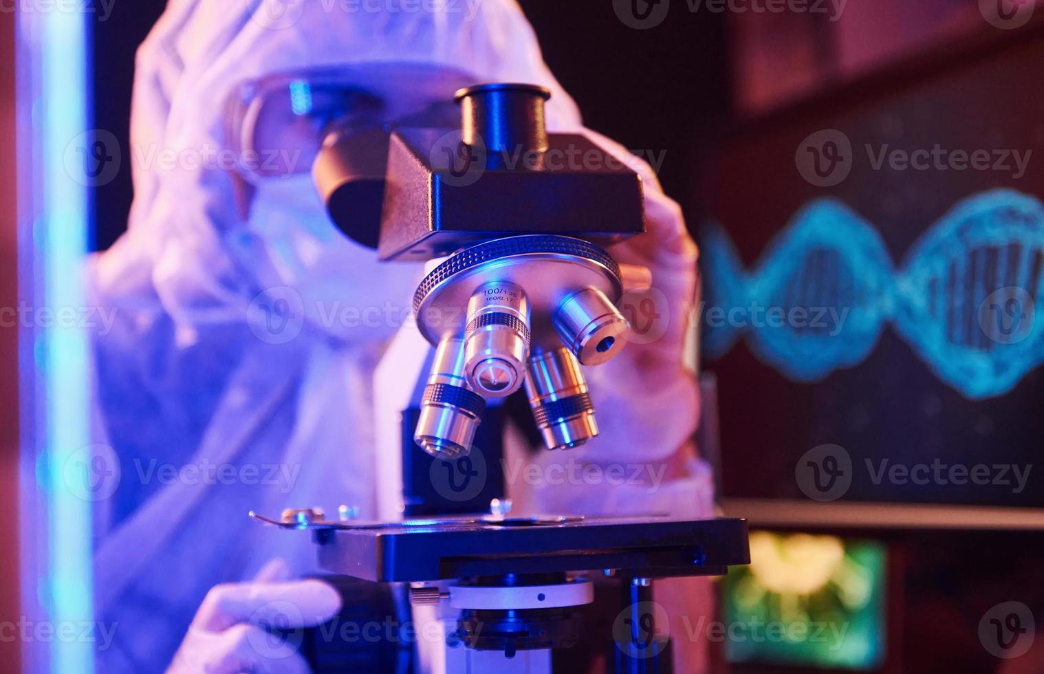 Nurse in mask and white uniform sitting in neon lighted laboratory with computer, microscope and medical equipment searching for Coronavirus vaccine photo