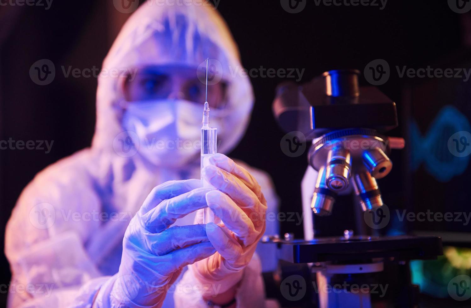 Nurse in mask and white uniform, holding syringe and sitting in neon lighted laboratory with computer and medical equipment searching for Coronavirus vaccine photo