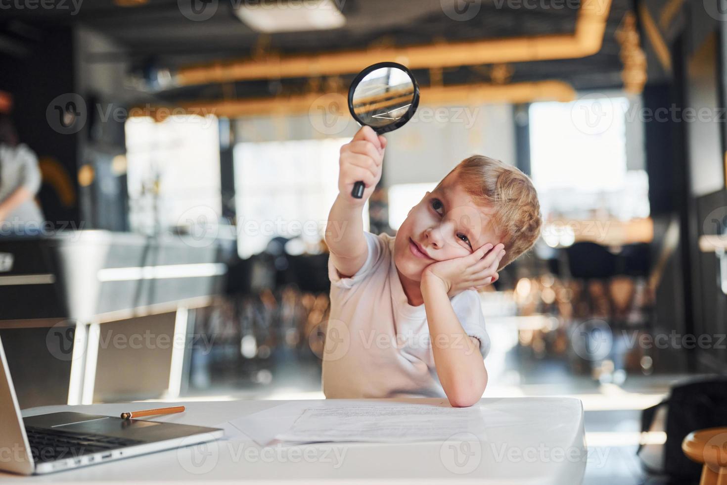 Smart child in casual clothes with laptop on table have fun with magnifying glass photo