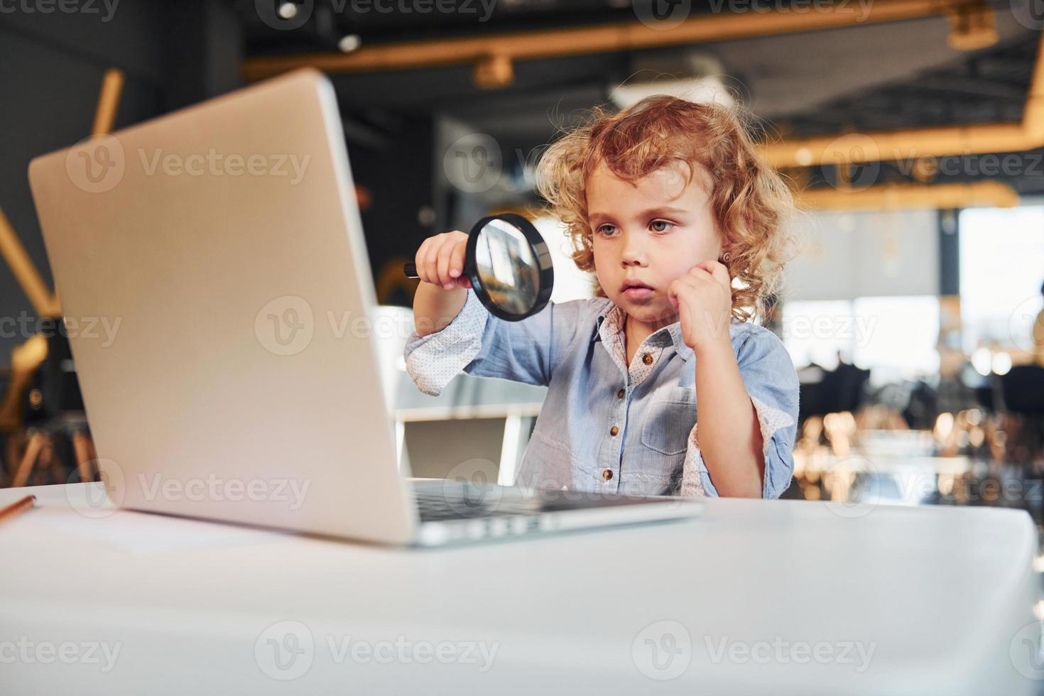 Smart child in casual clothes with laptop on table have fun with magnifying glass photo