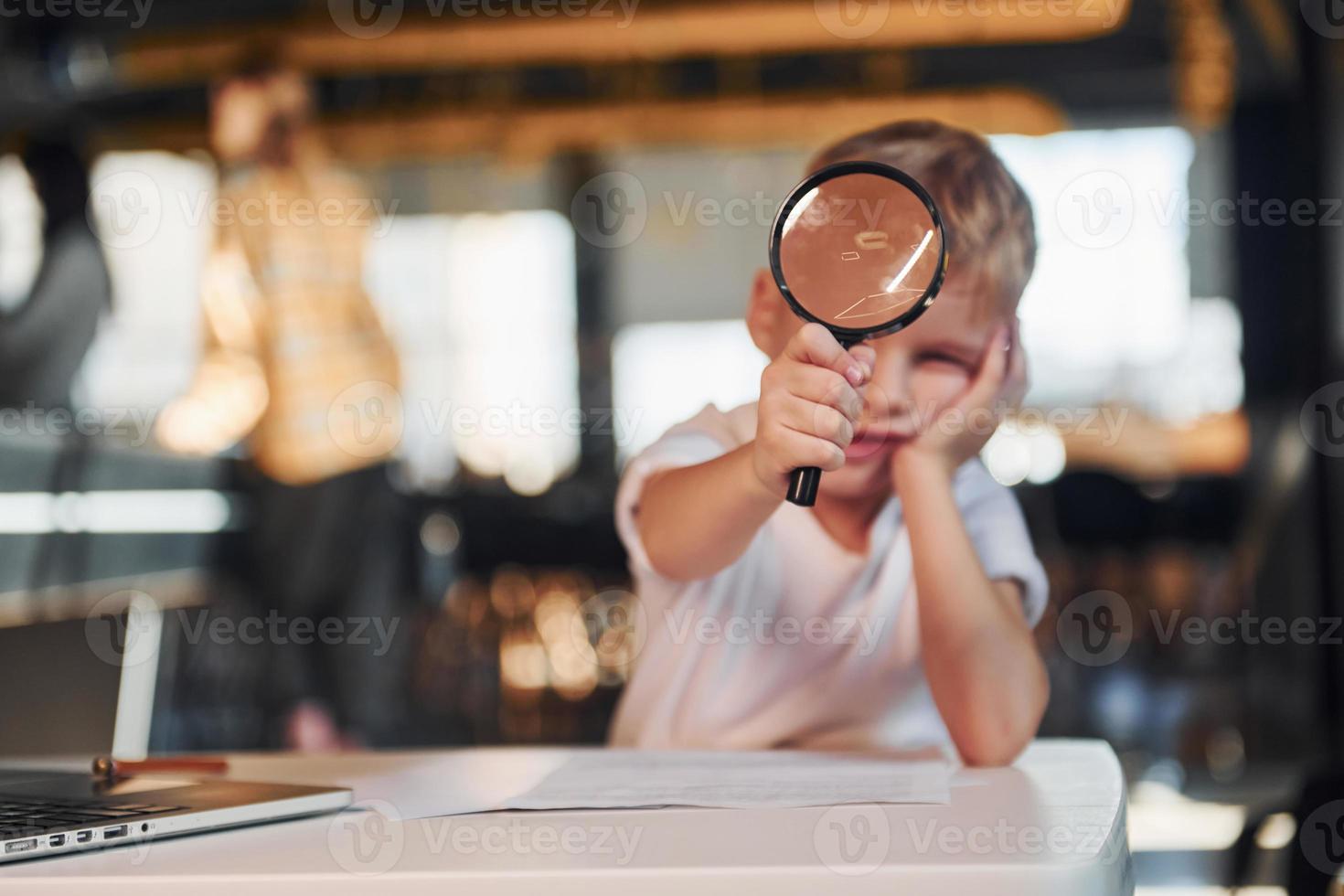 Smart child in casual clothes with laptop on table have fun with magnifying glass photo