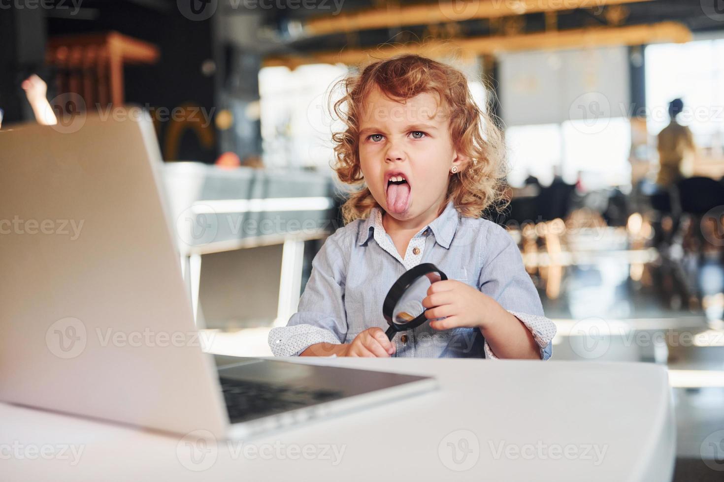 Smart child in casual clothes with laptop on table have fun with magnifying glass photo