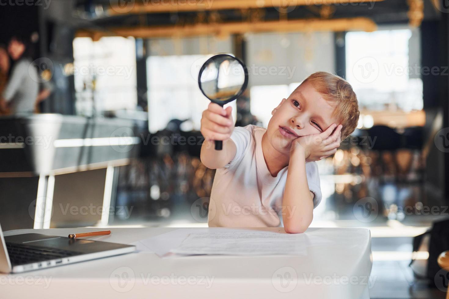 Smart child in casual clothes with laptop on table have fun with magnifying glass photo