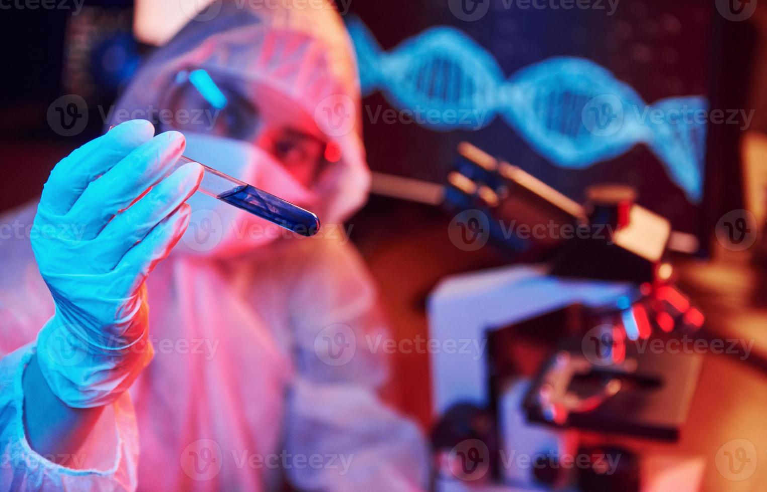 Nurse in mask and white uniform, holding tube with liquid and sitting in neon lighted laboratory with computer and medical equipment searching for Coronavirus vaccine photo