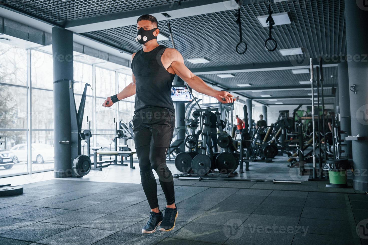 Strong young man in sportive clothes and scary mask doing exercise by using jump rope in gym photo