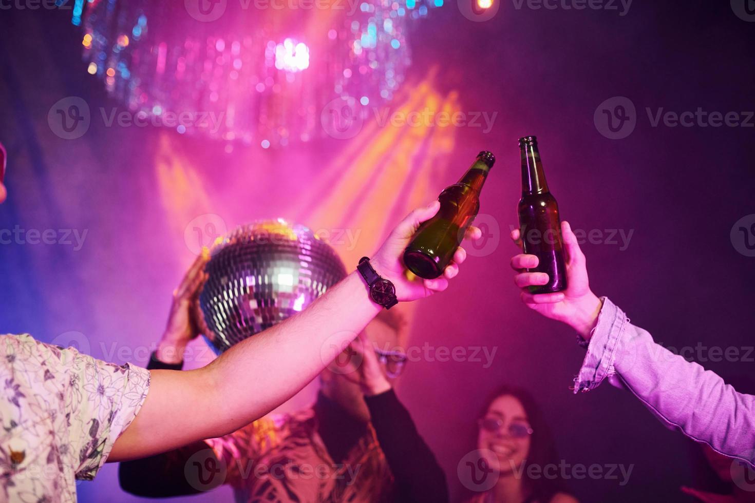 Celebrating and knocking bottles with alcohol. Young people is having fun in night club with colorful laser lights photo