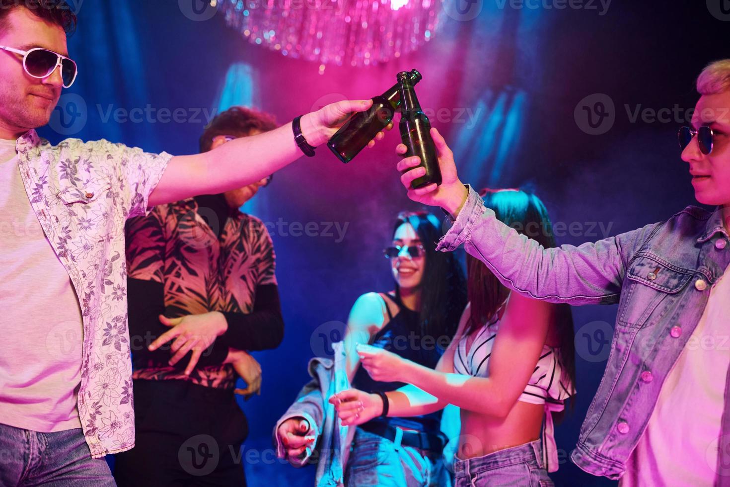 Celebrating and knocking bottles with alcohol. Young people is having fun in night club with colorful laser lights photo