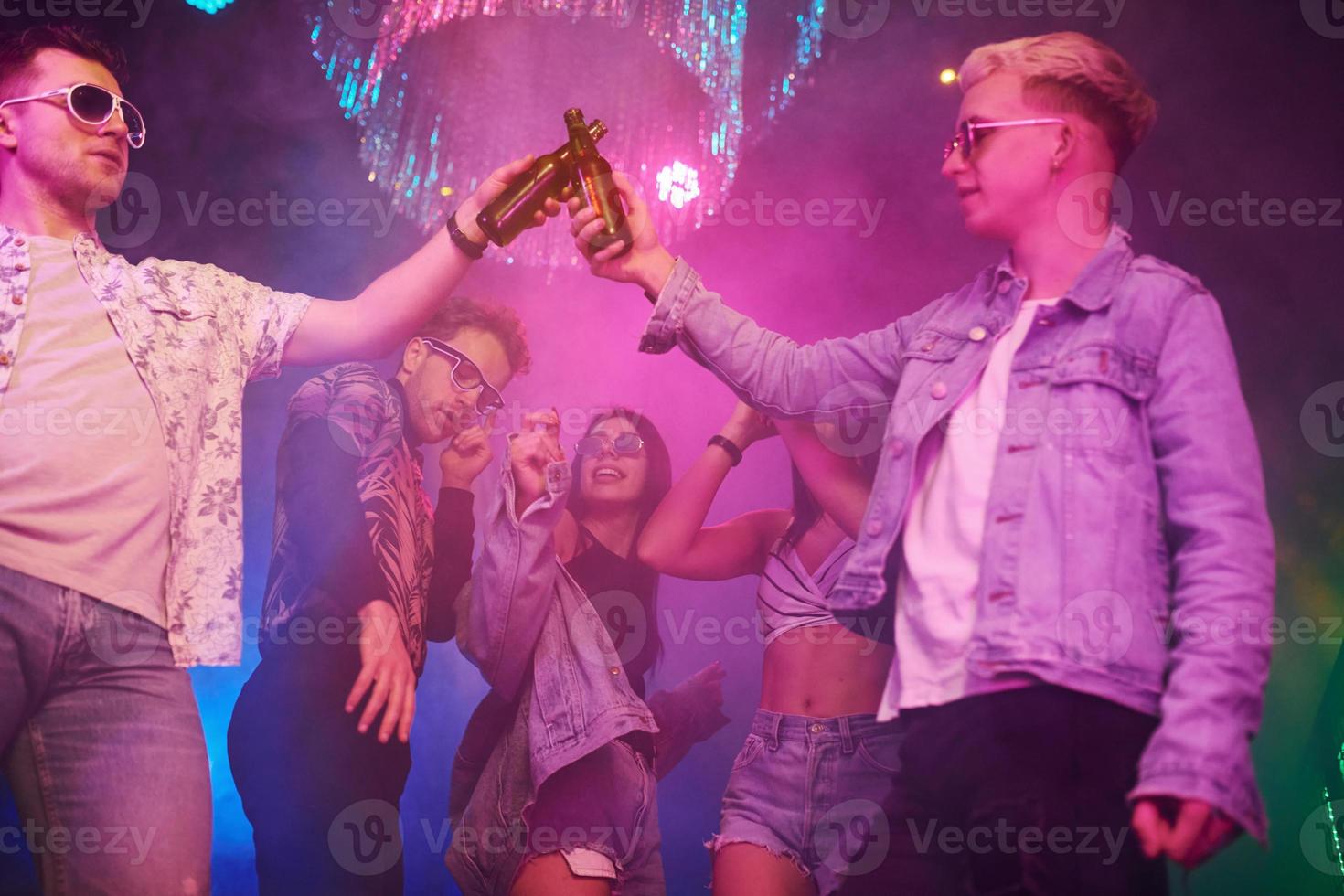 Celebrating and knocking bottles with alcohol. Young people is having fun in night club with colorful laser lights photo