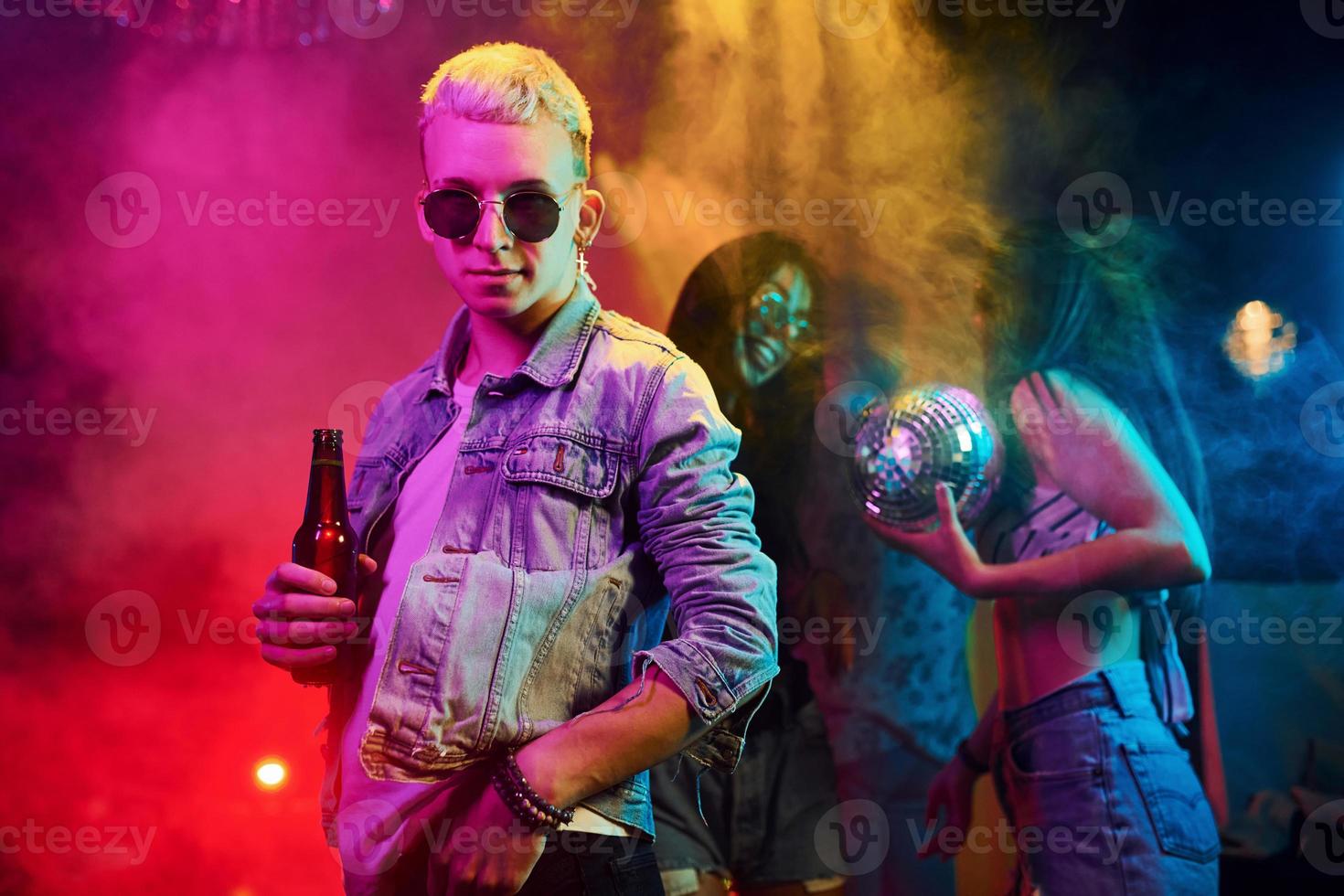 Hipster guy in sunglasses and with bottle of alcohol posing for camera in front of young people that having fun in night club with colorful laser lights photo