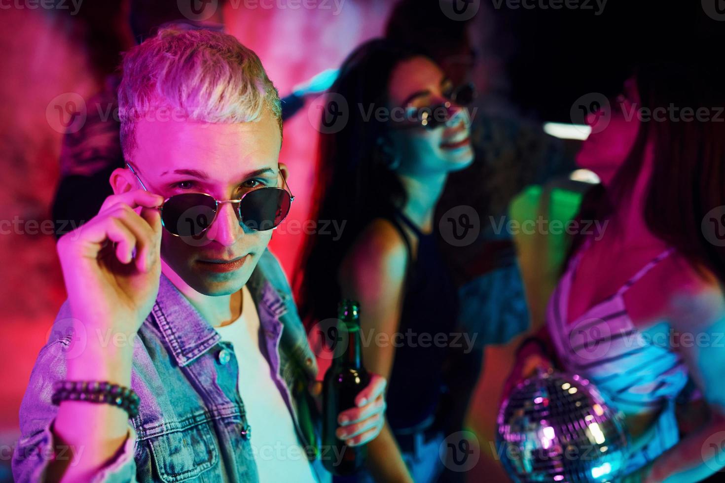 Hipster guy in sunglasses and with bottle of alcohol posing for camera in front of young people that having fun in night club with colorful laser lights photo